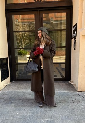 Mujer con abrigo y pantalones marrón oscuro, gorro gris, guantes rojos y bolso negro, de pie en la acera cerca de puertas de cristal y un cartel de recepción.