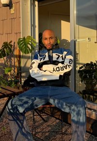 Melna ādas jaka ar baltiem un ziliem akcentiem, kas rotāta ar plāksteriem. Zilas džinsa bikses. Fonā augi un saules gaisma. Teksturēta virsma apakšā.