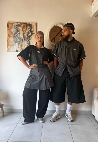 Dos personas están de pie una al lado de la otra. La mujer lleva una camiseta de malla negra, una falda gris plisada y unos pantalones anchos negros. El hombre viste una chaqueta negra y pantalones cortos.