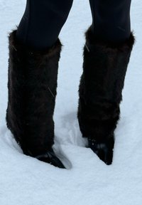 Ciorapi de blană artificială negri, purtați peste pantofi negri, pe un fundal de zăpadă albă. Blana este densă, cu o textură moale și lungimi inegale.