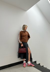 Pulover Adidas maroniu cu dungi albe, fustă în carouri gri cu o despicătură laterală, adidași roz și o geantă bordeaux cu accente argintii.