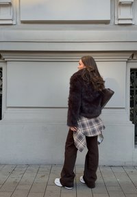 Femme aux longs cheveux portant une veste en fourrure sombre, une chemise à carreaux nouée autour de la taille, un pantalon foncé et des baskets, marchant sur une rue pavée à côté d'un mur en pierre.