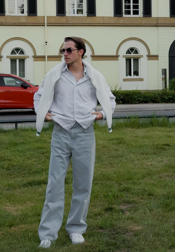 Pessoa vestindo um suéter branco canelado, uma camisa de botão listrada, jeans cinza claro e tênis brancos. Em pé sobre a grama com um edifício ao fundo.