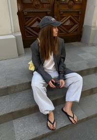 Szary, oversize'owy żakiet, biały t-shirt, szare dresy, czarne klapki, żółta torba, siedzący na kamiennych schodach, trzymający okulary.