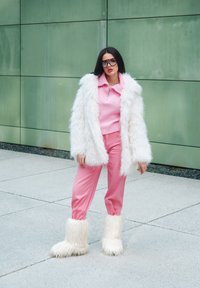 Růžový pletený top s límečkem, bílý kabát z umělé kožešiny, růžové kalhoty a chlupaté bílé boty stojící na dlážděném povrchu před zelenou zdí.