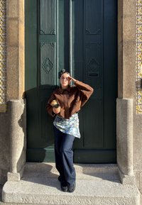 Kvinde iført brun jakke og jeans står på stentrapper foran en stor grøn dør, holder en kaffekop og justerer solbriller.