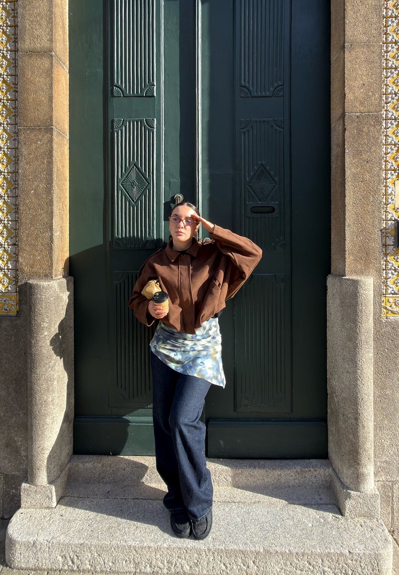 Femme portant une veste marron et un jean, debout sur des marches en pierre devant une grande porte verte, tenant une tasse de café et ajustant ses lunettes de soleil.