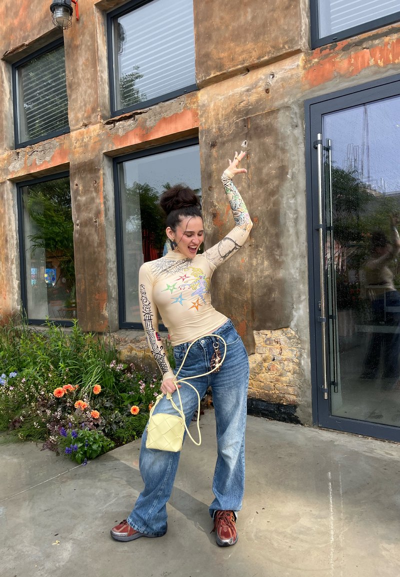 Beige Langarmoberteil mit bunten Grafikdrucken, kombiniert mit oversized blauen Jeans. Die Accessoires umfassen eine kleine gelbe Tasche und braune Schuhe.