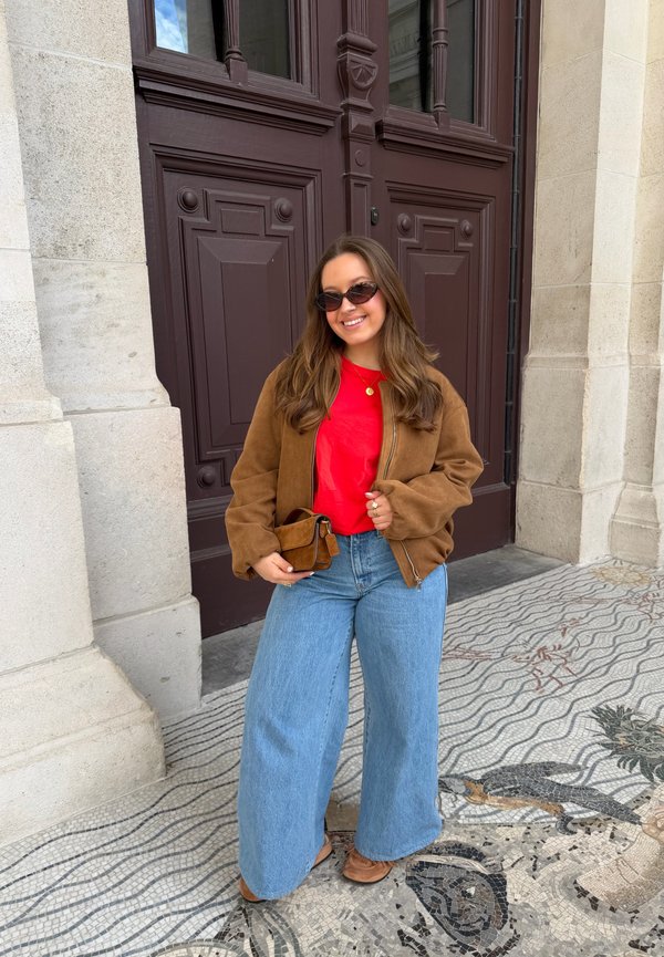 Young woman wearing sunglasses, brown jacket, red shirt, and wide blue jeans, standing on mosaic floor in front of large purple wooden door.