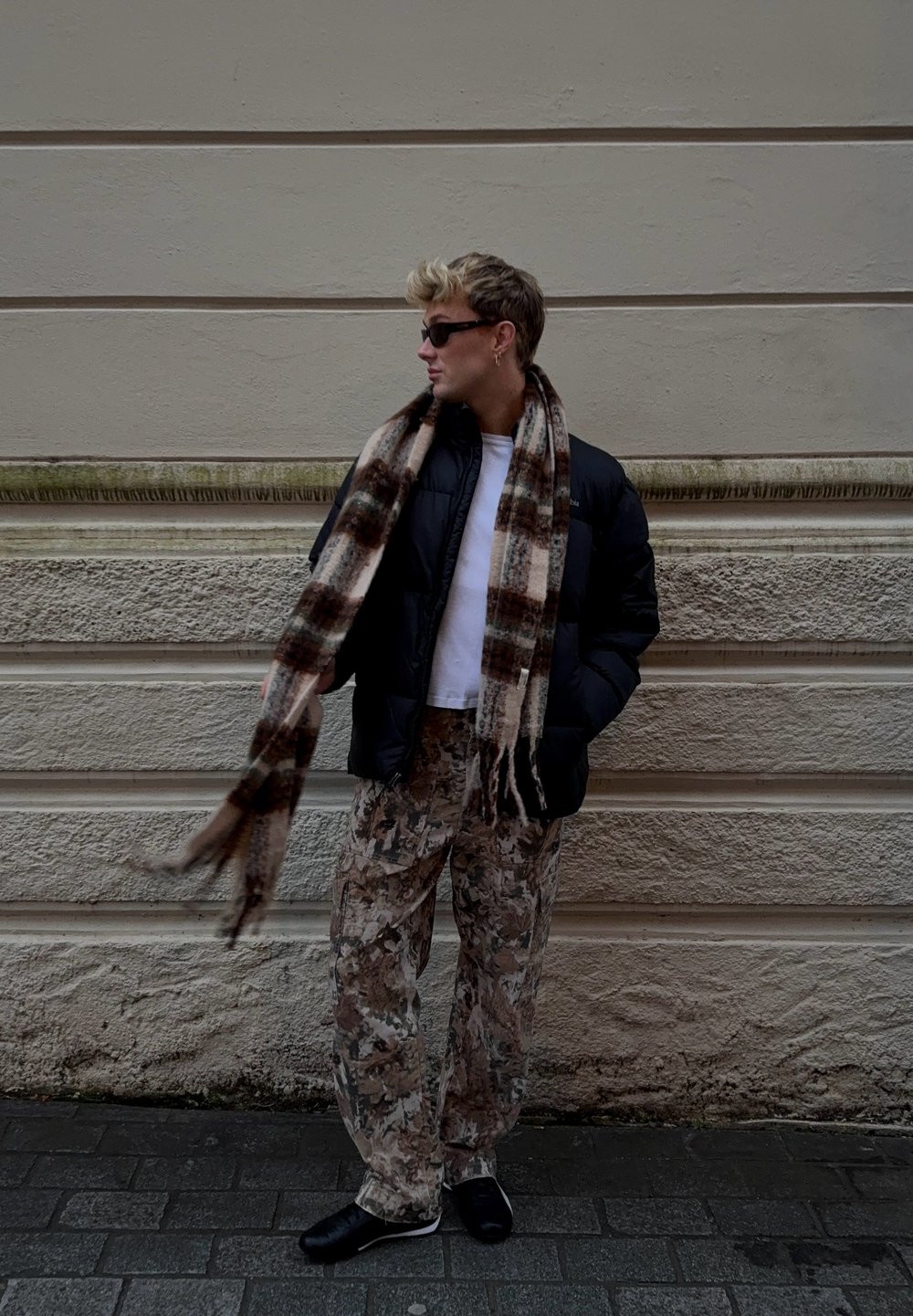 Casaco puffer preto, camisa branca, cachecol castanho com padrão, calças camufladas bege, sapatos pretos. De pé contra uma parede texturizada bege no pavimento.