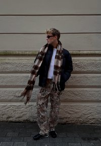 Casaco puffer preto, camisa branca, cachecol castanho com padrão, calças camufladas bege, sapatos pretos. De pé contra uma parede texturizada bege no pavimento.