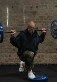 Αθλητής σε ναυτικό hoodie και μοτίβο πράσινες κολάν εκτελεί ένα λούγκα με μια μπάρα, που στηρίζεται σε μια μπλε πλακέτα βάρους στο πάτωμα.