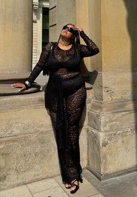 Completo in pizzo nero con maniche lunghe e uno spacco sulla coscia, caratterizzato da un corpetto aderente e un soprabito fluente, abbinato a tacchi neri.