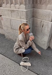Lichtgrijze gestreepte oversized blazer, blauwe jeans, bruine laarzen en een witte handtas. Persoon houdt een doorzichtige beker vast, zittend bij een gestructureerde muur.
