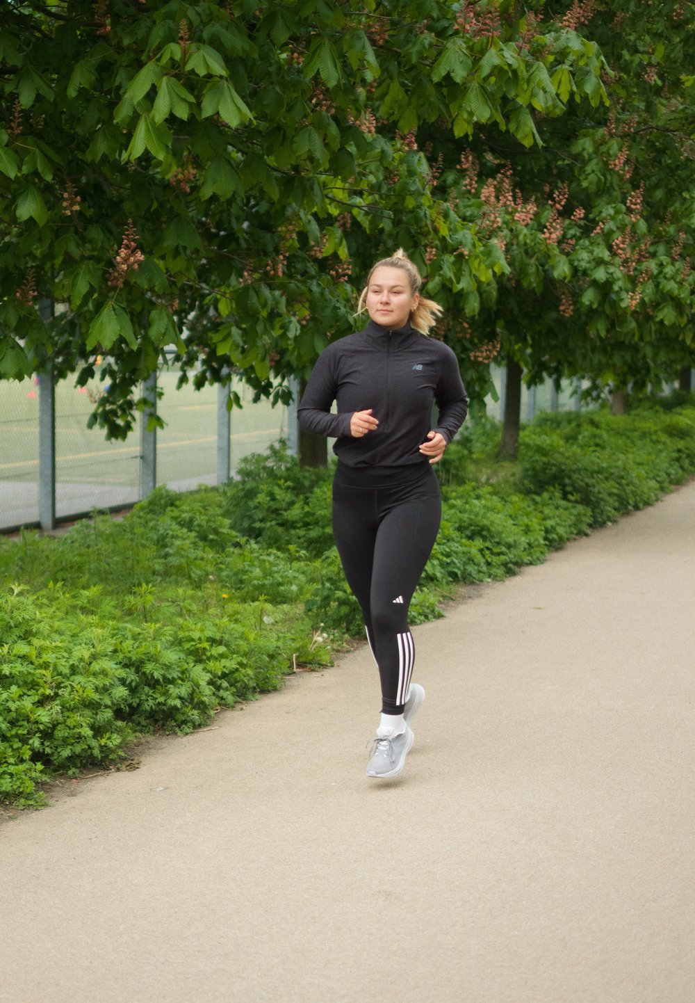 Hardloper in een zwarte ritsjack en zwarte leggings met witte strepen, met grijze sportschoenen, joggend op een pad omringd door groen.