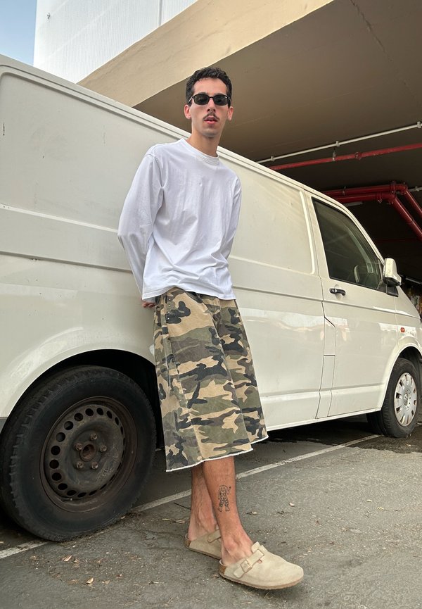 Homme portant des lunettes de soleil, une chemise blanche à manches longues, un short camouflage et des chaussures beiges sans lacets, appuyé contre une camionnette blanche dans un parking.