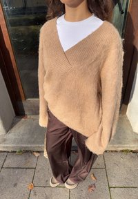 Lichtbruine, oversized trui met V-hals, gemaakt van zacht materiaal, gecombineerd met donkerbruine, losse broek en crèmekleurige instapschoenen.