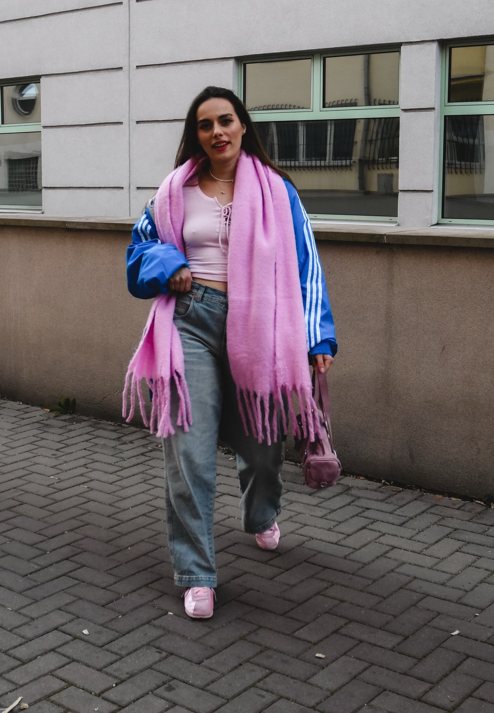 Μπλε τζάκετ Adidas, ροζ υπερμεγέθη κασκόλ, ροζ κοντό τοπ, γκρι τζιν με ψηλή μέση, ροζ αθλητικά παπούτσια και μια μικρή ροζ τσάντα. Φόντο με πλακόστρωτο μονο πάτι.