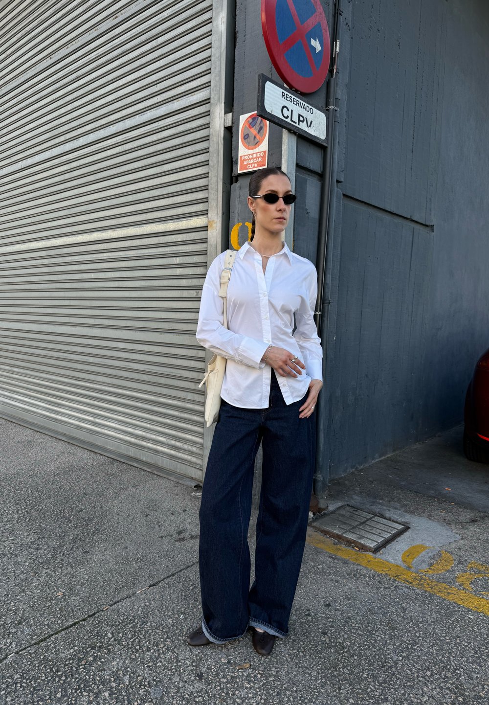 Biała koszula z guzikami, ciemne dżinsy o szerokich nogawkach, czarne okulary przeciwsłoneczne, srebrna biżuteria i jasna torba na ramię. Tło przedstawia metalową żaluzję.