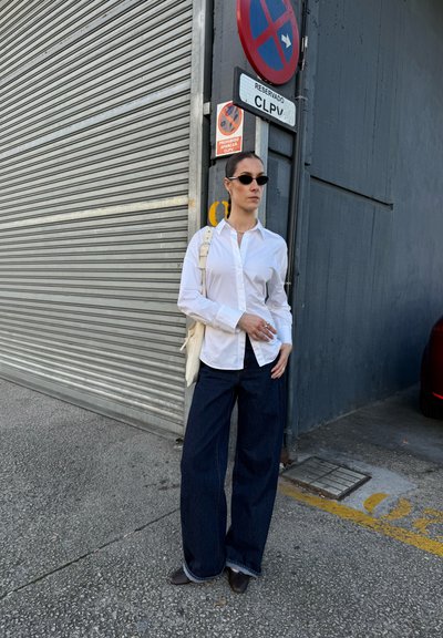Camisa blanca de botones, vaqueros rectos oscuros, gafas de sol negras, joyería plateada y un bolso de mano de color claro. El fondo presenta un shutter de metal.