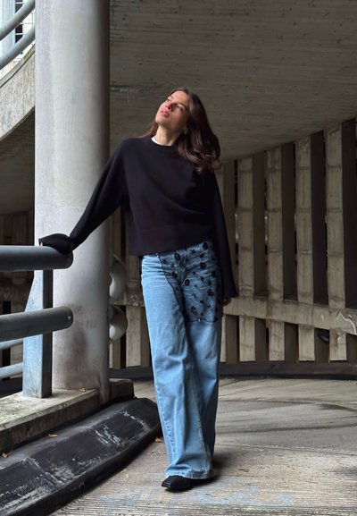 Mujer con suéter negro y jeans azules con un sobrepuesto floral negro, apoyada en una barandilla en un estacionamiento de concreto, mirando hacia arriba.