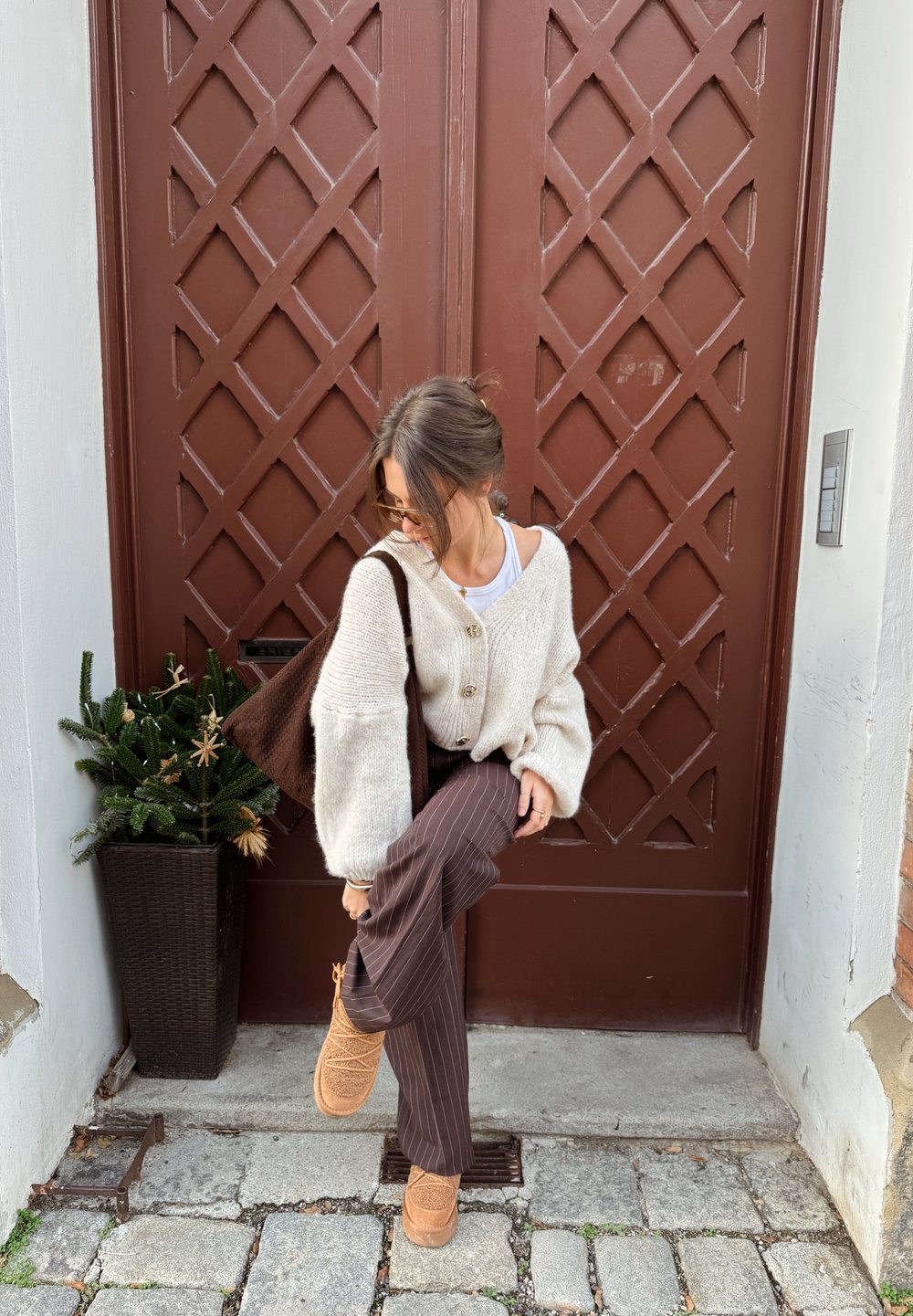 Cardigan en grosse maille crème avec de gros boutons, associé à un pantalon marron à rayures fines et des chaussures couleur cognac. Plante verte en pot à proximité, en arrière-plan une porte marron.