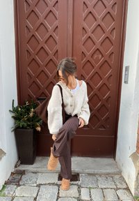 Cardigan en grosse maille crème avec de gros boutons, associé à un pantalon marron à rayures fines et des chaussures couleur cognac. Plante verte en pot à proximité, en arrière-plan une porte marron.