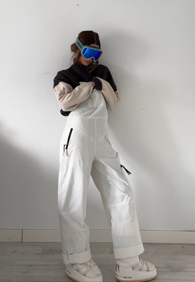 Overalls de nieve blancos y holgados con bolsillos con cremallera, combinados con una camiseta negra y beige y gafas de esquí azules, con una textura acogedora.