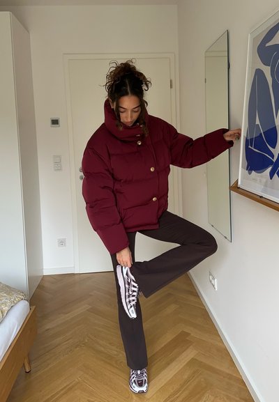 Chaqueta acolchada de color burdeos con cuello alto, presenta grandes botones. Pantalones marrones, zapatillas con estampado y acentos blancos, y un suelo de madera visible.