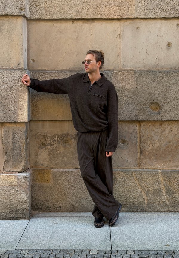 Hombre con un suéter oscuro de gran tamaño y pantalones a rayas se apoya en una pared de piedra, mirando hacia un lado con gafas de sol puestas.