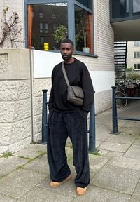 Juodas oversize džemperis, laisvi juodi kelnių, rudos spalvos avalynė ir tamsus pagalvėlės tipo krepšys. Tekstūruota medžiaga ir laisvas siluetas. Miestovaizdis.