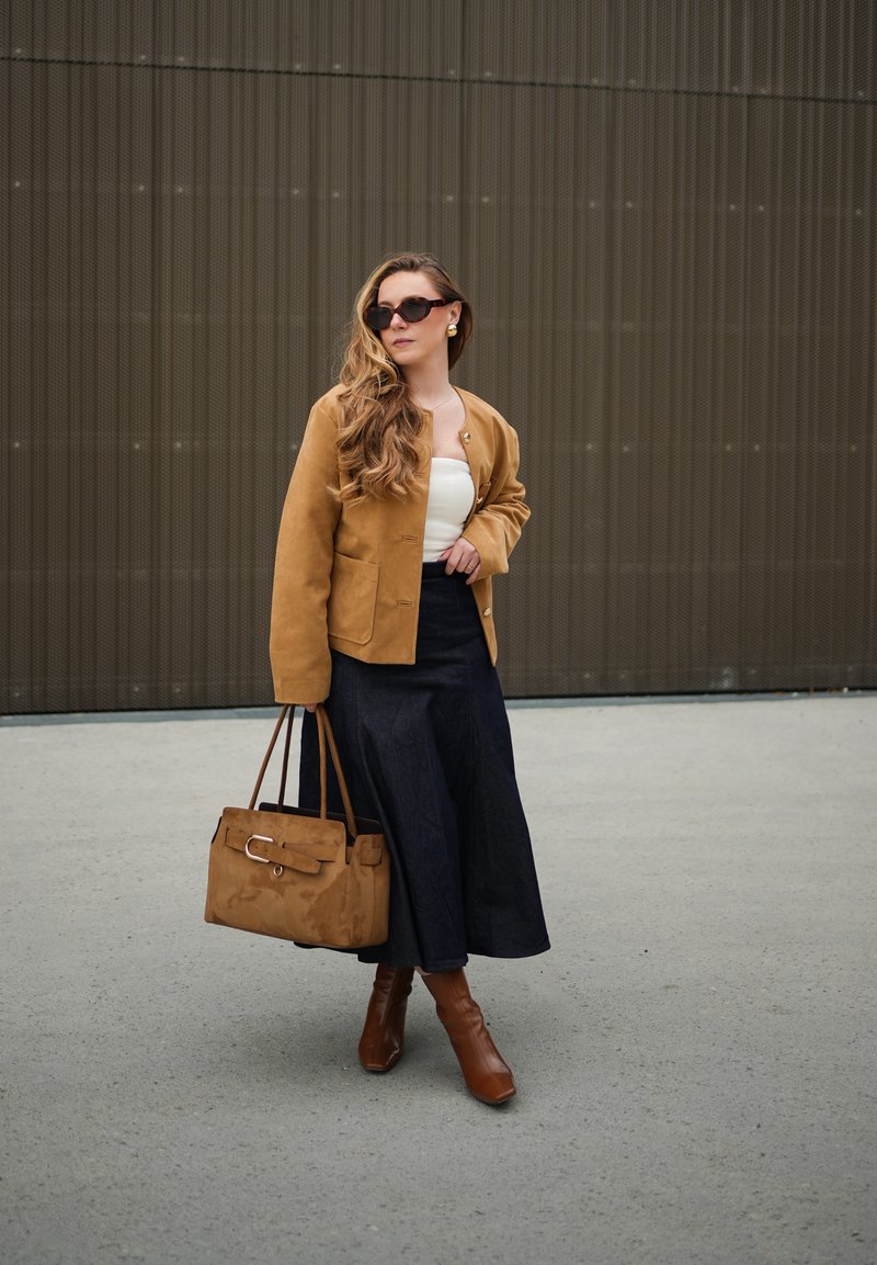 Femme aux longs cheveux ondulés portant des lunettes de soleil, une veste marron, un haut blanc, une jupe noire, des bottes marron, tenant un sac à main marron devant un mur texturé.