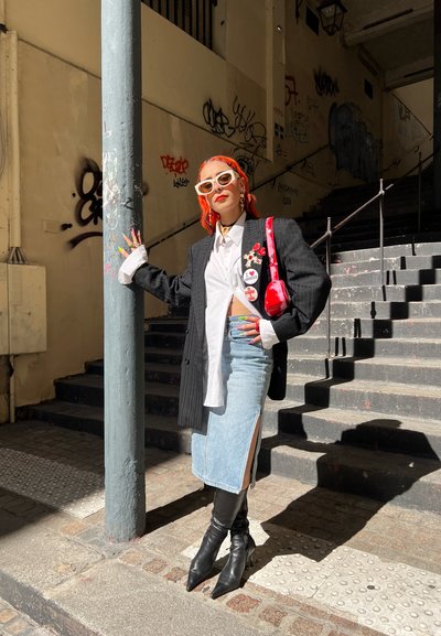 Blazer oversize a rayas negras, blusa blanca, falda de mezclilla con aberturas laterales, botas negras de puntera afilada, bolsa roja y gafas de sol blancas.