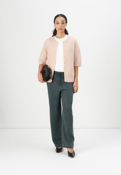 Cárdigan corto de manga corta en rosa claro, con botones sobre una camiseta blanca, combinado con pantalones vaqueros de pierna ancha en verde oscuro y un bolso de mano negro.