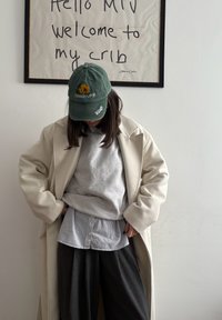 Personne portant un manteau beige, un pull gris, une chemise rayée, un pantalon foncé et une casquette verte avec le texte "TORREMOLINOS Málaga", se tenant sous un panneau encadré "Hello MTV".