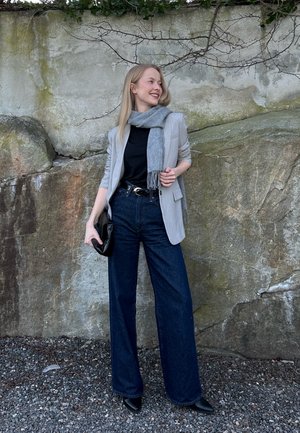 Mujer de pie junto a una pared de rocas cubiertas de musgo, vestida con blazer gris, blusa negra, pantalones vaqueros de pierna ancha, bufanda gris, botas negras, sosteniendo un bolso de mano negro, sonriendo hacia la izquierda.