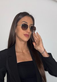 Óculos de sol com lentes redondas e escuras, e detalhes metálicos dourados. A armação é fina e brilhante, combinando com uma roupa preta e cabelo comprido e liso.