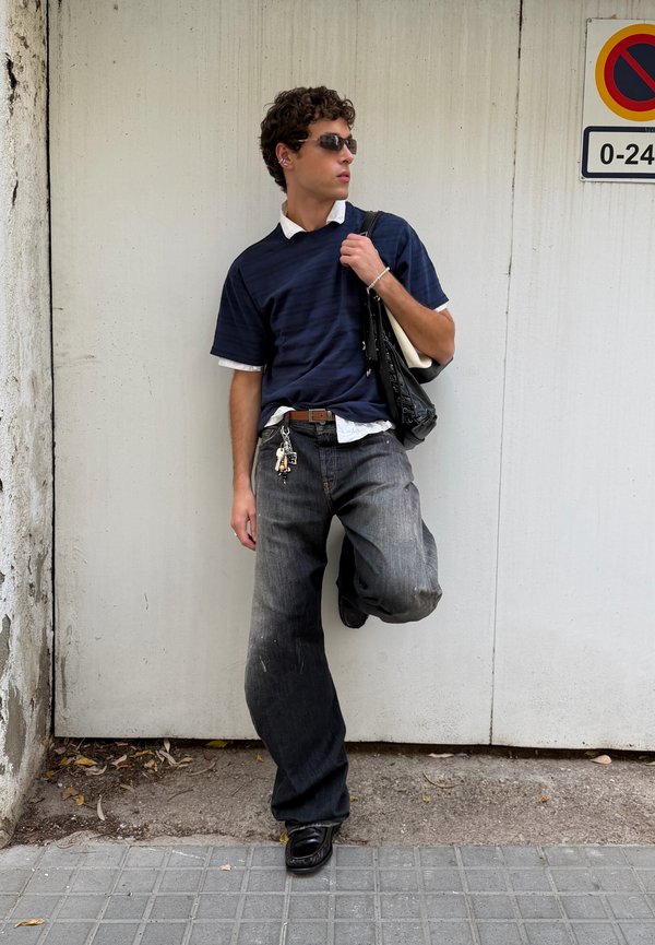 Joven con gafas de sol, camisa azul marino y jeans negros holgados, apoyado contra una pared blanca cerca de una señal de prohibido aparcar, sosteniendo una bolsa negra.