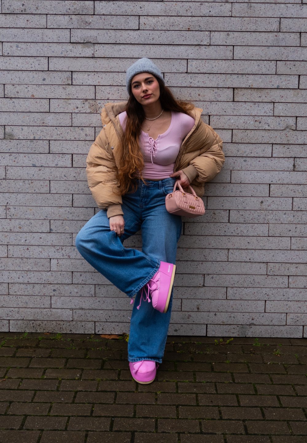 Beige puffer jacket, light pink top, high-waisted blue jeans, bright pink platform trainers, and a small pink handbag, against a brick wall.