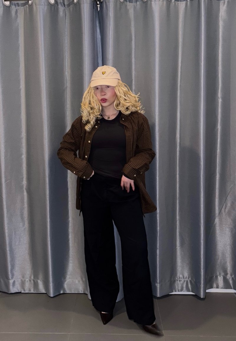 Brun ternet oversized skjorte, sort top, sorte bukser med vid ben og en beige baseballkasket. Krøllet blondt hår, står foran grå gardiner.