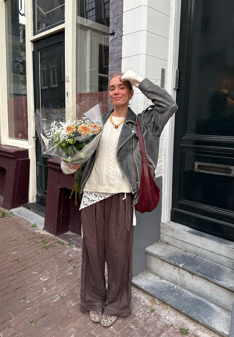Vrouw in leren jasje met een boeket bloemen, staande op een stenen stoep nabij een gebouwingang met een donkergroene deur en trappen.