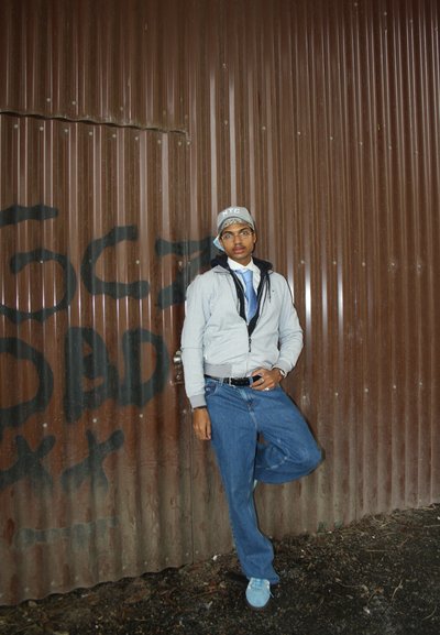 Chaqueta gris con cremallera sobre una camisa blanca y una corbata azul, combinada con unos jeans azules holgados y unas zapatillas ligeras azules, frente a una pared de cartón corrugado marrón.