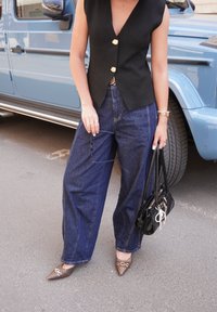 Gilet sans manches noir avec des boutons dorés, jean bleu à jambes larges, chaussures brunes à bout pointu avec un motif, et un sac à main noir.