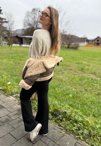 Beige og brun pelsforet jakke med en blød tekstur, kombineret med sorte vide bukser og hvide sneakers. Udendørs græsbevokset baggrund.