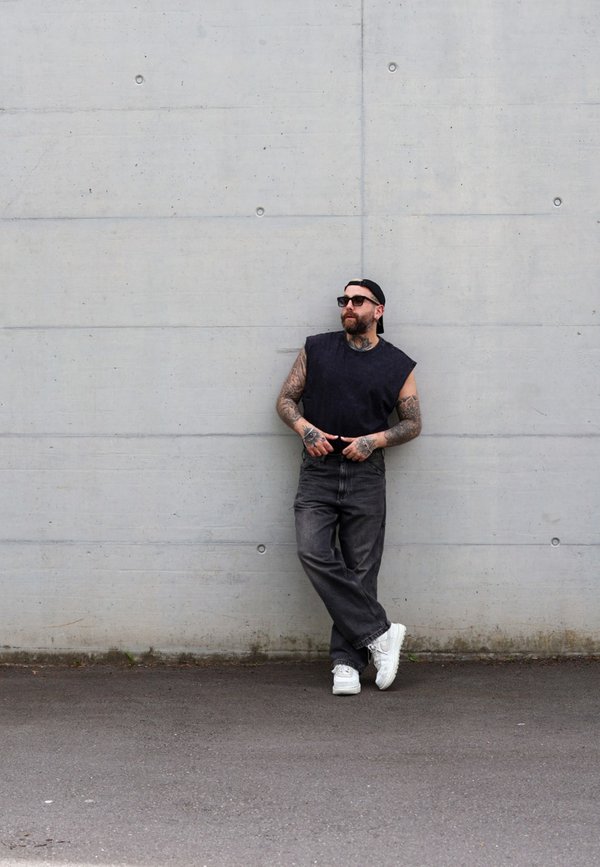 Maglietta nera senza maniche, jeans scuri, sneakers bianche. Tatuaggi sulle braccia, cappellino nero, occhiali da sole. Appoggiato a un muro di cemento grigio.