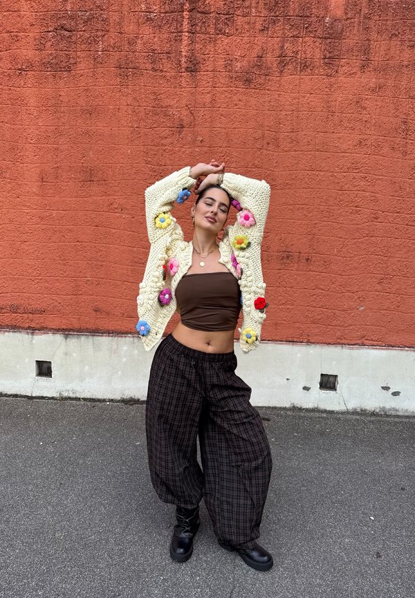 Vrouw in kleurrijk gebreide cardigan en losse broek staat met armen omhoog tegen een gestructureerde oranje muur buiten.