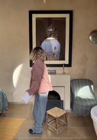 Skjorte i ternet orange og hvid, grå bukser. Teksturerede vægge, stengulv, vævet skammel, rundt spejl og indrammet kunstværk i baggrunden.