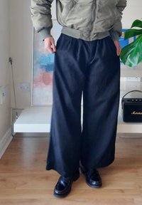 Persoon met zwarte wijde broek, zwarte schoenen en een groene bomberjack die op een houten vloer binnen staat.