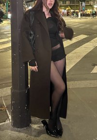 Casaco castanho longo sobre uma blusa preta, calções de renda e ligas pretas. Saltos altos pretos brilhantes. A pessoa está de pé ao lado de um poste numa esquina à noite.