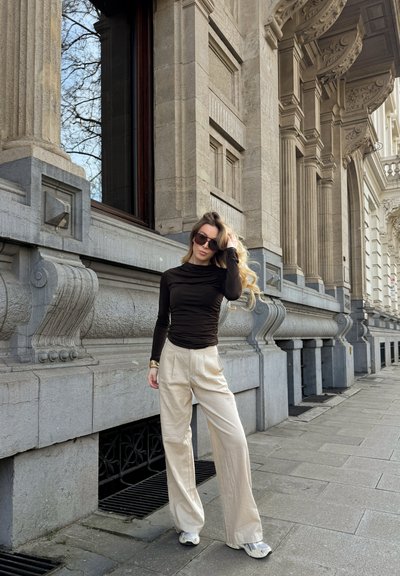 Top de manga larga de color marrón oscuro combinado con pantalones sueltos de color beige claro. Gafas de sol y zapatillas deportivas complementan el atuendo casual. Fondo texturizado.